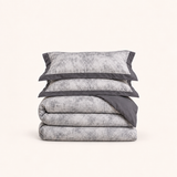 Noctua Paris — Parure de lit en gaze de coton gris, deux taies à bordure unie et housse pliée, sur fond ivoire.